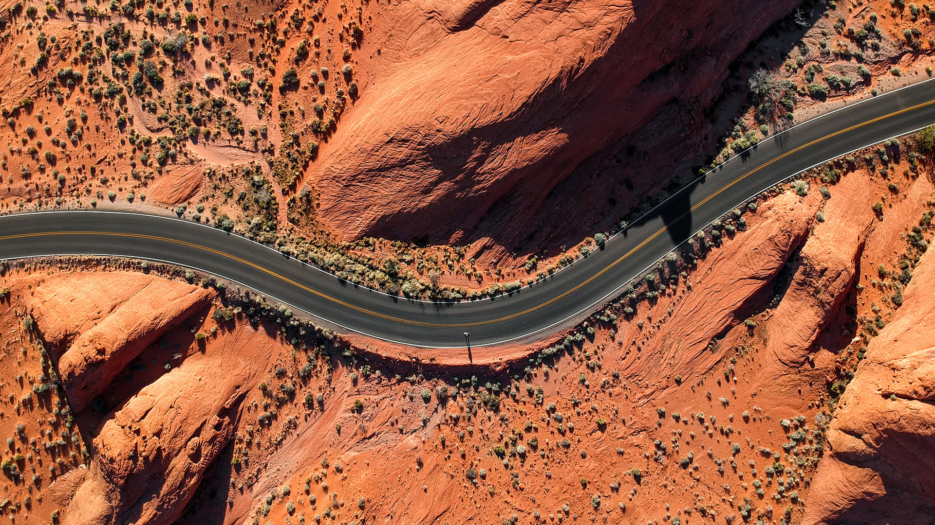 Aerial view of a winding road cutting through a desert landscape with reddish-brown rock formations and sparse vegetation.
