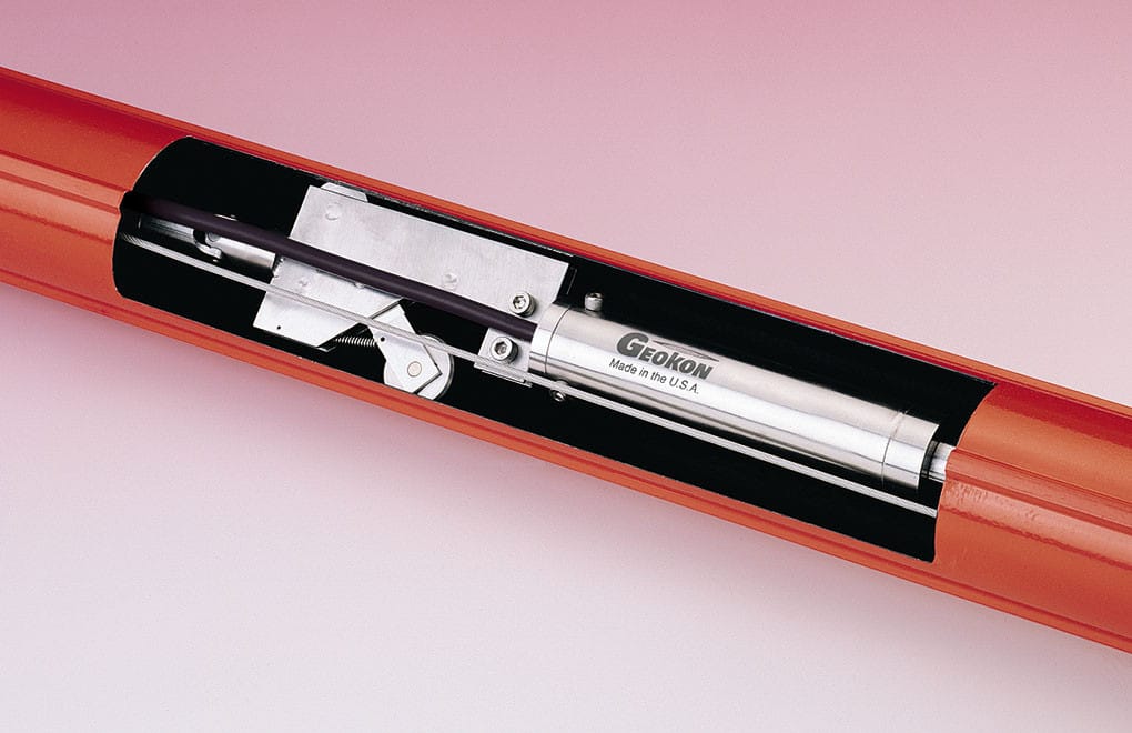 A close-up of a Geokon instrument installed inside a cutaway section of a red cylindrical pipe, revealing internal components.