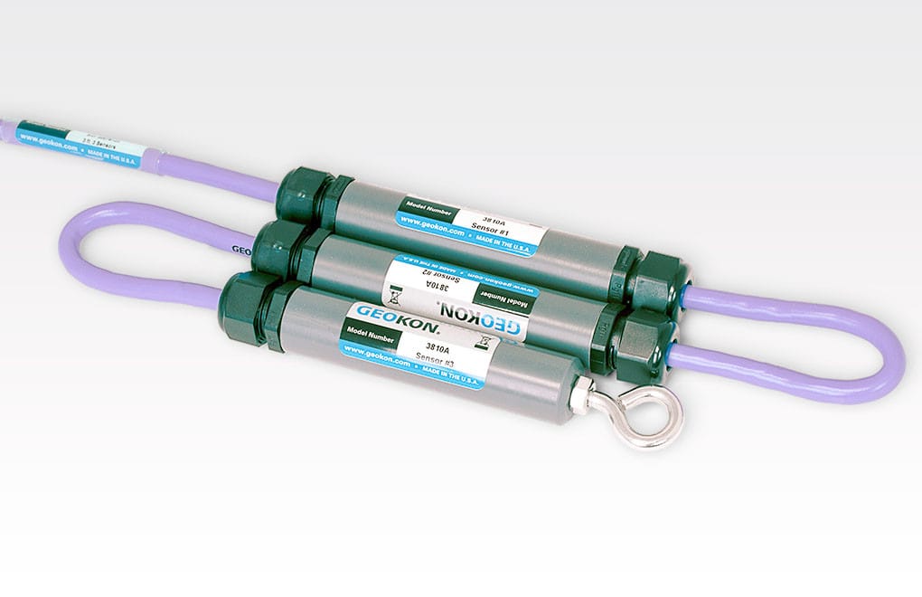 Two cylindrical metallic sensors with attached purple cables and labels, one featuring a metal hook at the end, laid against a light background.