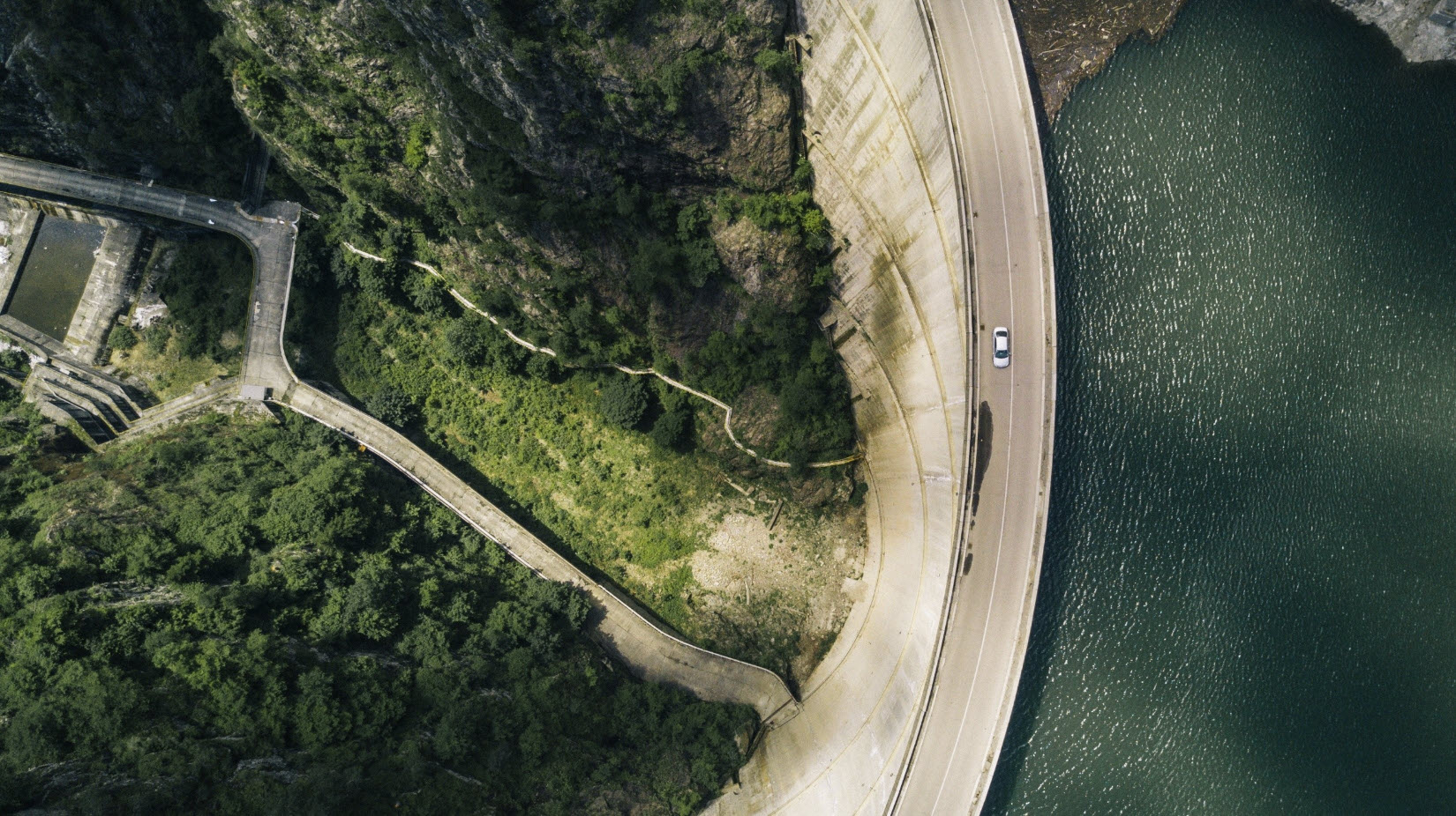 Aerial view of a car driving on a curved dam road, showcasing impressive geotechnical construction with water on one side and lush green vegetation on the other.