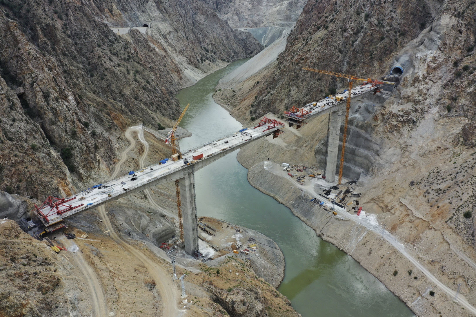 A partially constructed bridge spans a river between two rocky cliffs, with cranes and construction equipment visible at the site, showcasing impressive construction and geotechnical efforts.