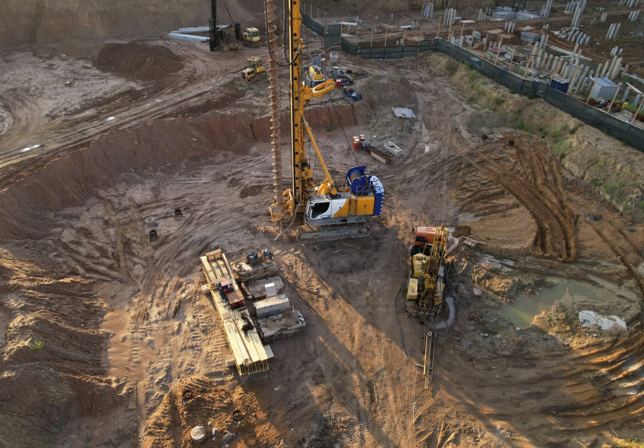 A large geotechnical drilling machine operates at a construction site with dirt piles, building materials, and equipment scattered around an excavated area.