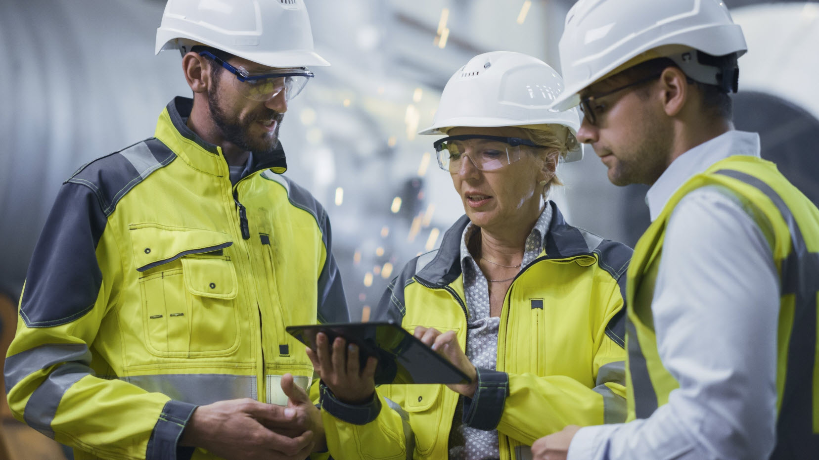 Three workers in safety gear and hard hats review geotechnical design information on a tablet in an industrial setting, with sparks visible in the background.