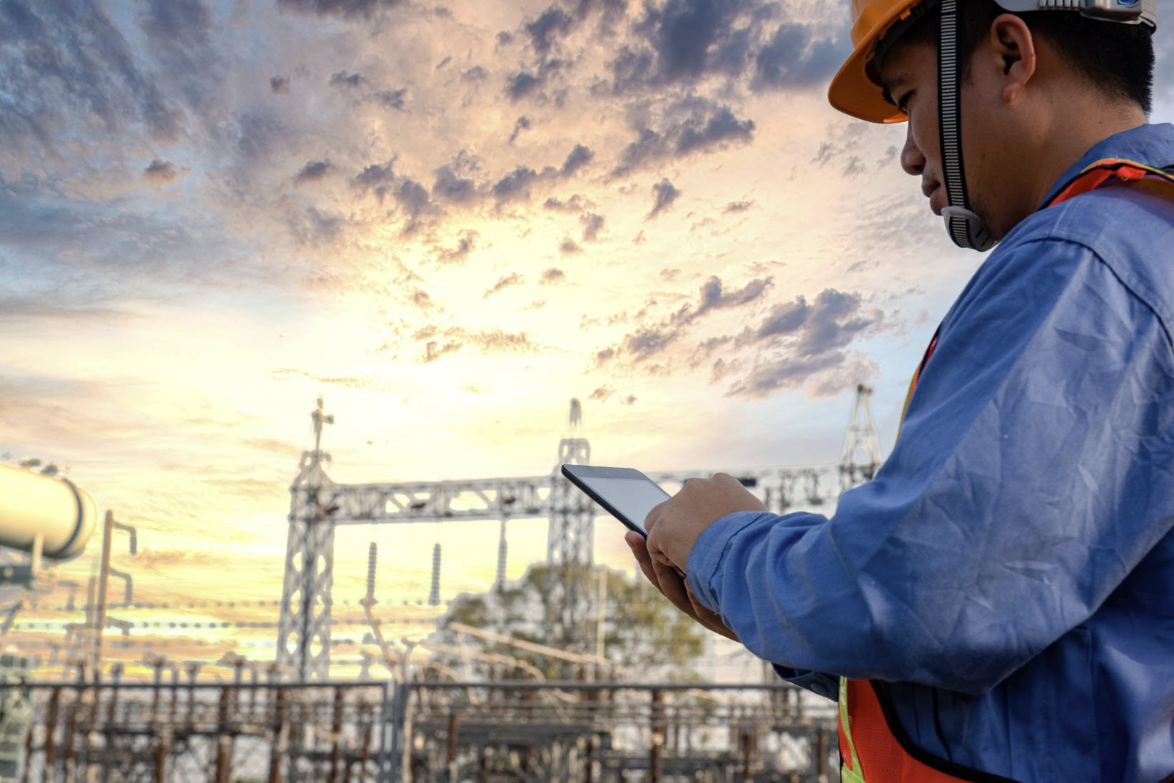 A worker wearing a hard hat and safety vest uses a tablet to monitor operations at an outdoor industrial facility with geotechnical and electrical infrastructure at sunset.