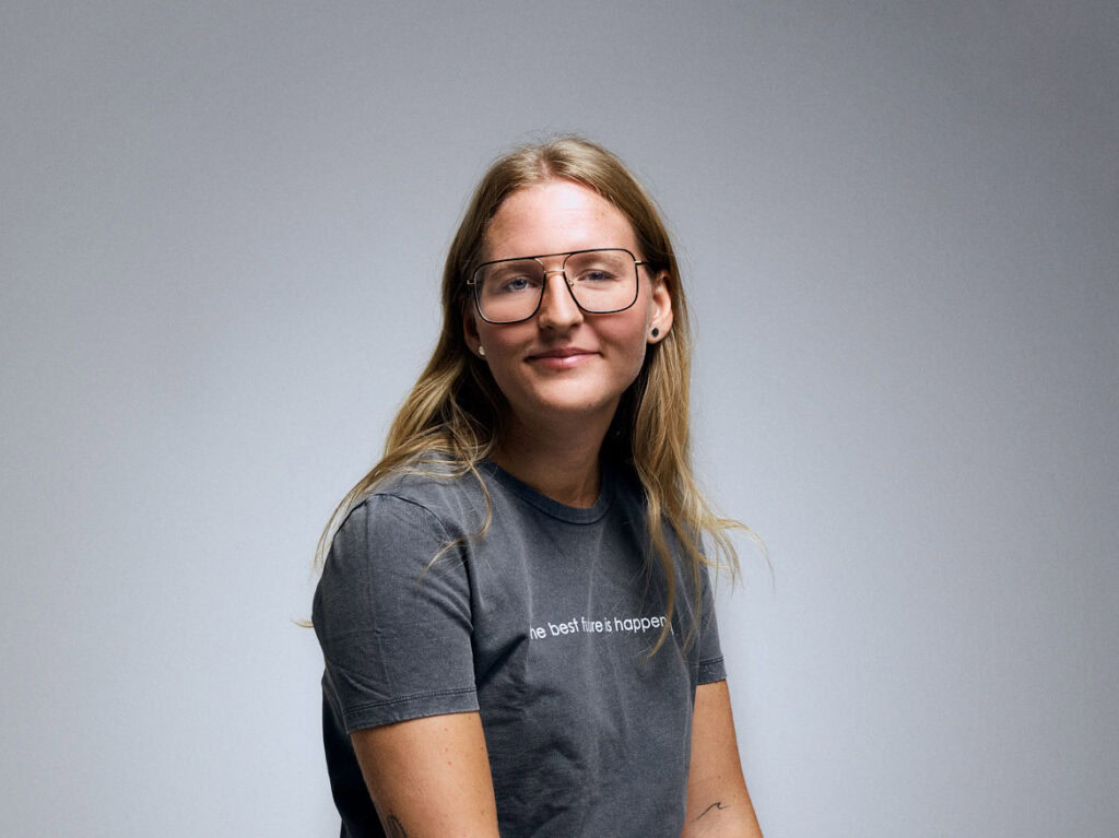 A woman with long blonde hair wearing glasses and a gray t-shirt sits against a plain light gray background, looking slightly to the side and smiling gently.