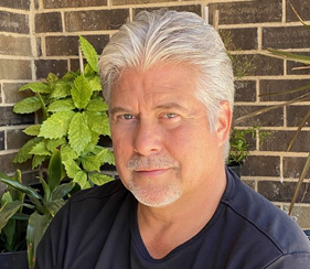Middle-aged man with silver hair and a black shirt, looking at the camera, attending a webinar, standing in front of a brick wall with green plants.