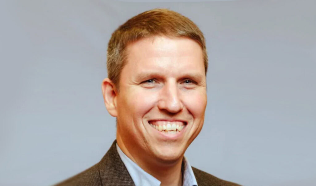 A man with short brown hair and a suit jacket smiles in front of a plain, light-colored background.