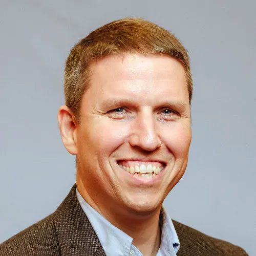 Man with short light brown hair wearing a brown blazer and light blue collared shirt, smiling in front of a plain neutral background, representing the positive outlook on America infrastructure and recent $1.2 trillion infrastructure funding.