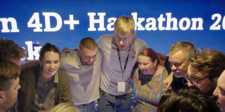 A group of people stand in a huddle with arms around each other in front of a blue screen displaying “Hackathon 4D+,” celebrating innovations in 4D modeling with Synchro+ software A group of people stand in a huddle with arms around each other in front of a blue screen displaying “Hackathon 4D+,” celebrating innovations in 4D modeling with Synchro+ software.