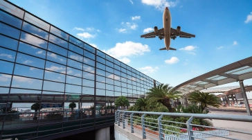 A commercial airplane flies overhead near a modern airport terminal with glass windows, palm trees, and blue sky.