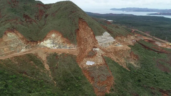 A large landslide cuts through a hillside, exposing soil and rocks and damaging the roadway; some construction materials are visible near the slide, along with what appear to be New Caledonian hatch patterns in the soil layers.