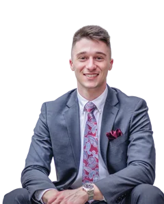 A smiling man in a gray suit with a pink tie and pocket square sits casually at an open site event.