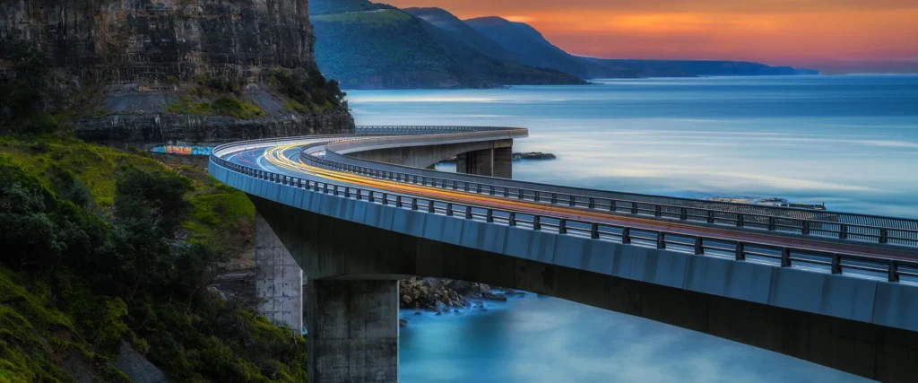 A curving bridge extends over the ocean at sunset, with cliffs on one side and blurred light trails from vehicles on the road.