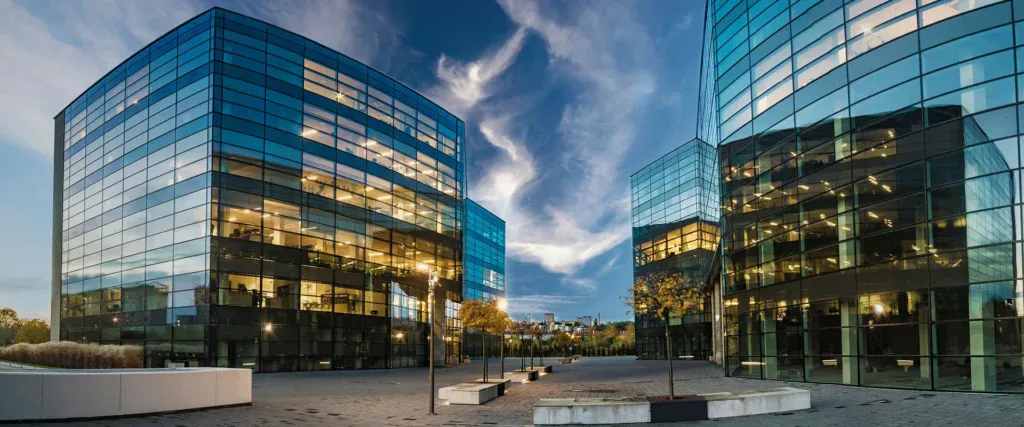 Modern glass office buildings with illuminated windows surround a paved courtyard at dusk, reflecting the evening sky.