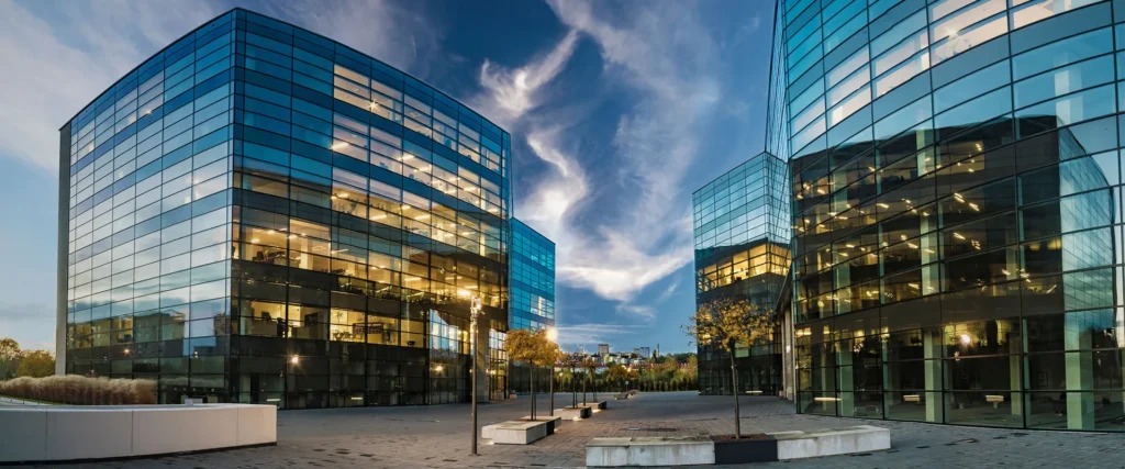 Modern office buildings with glass facades reflecting the sky, illuminated from within, surrounding an empty paved courtyard at dusk.