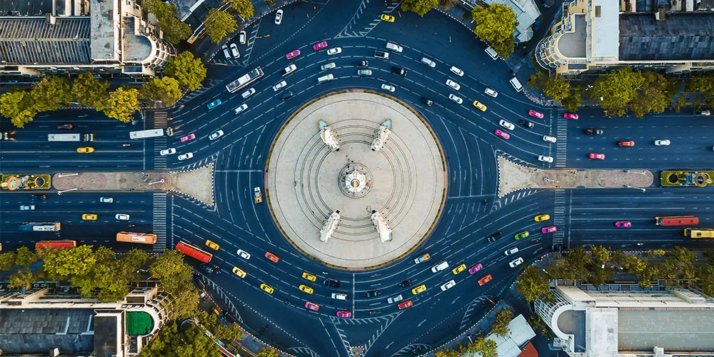 Aerial view of a large roundabout with a central monument, surrounded by roads with moving cars and bordered by trees and buildings.