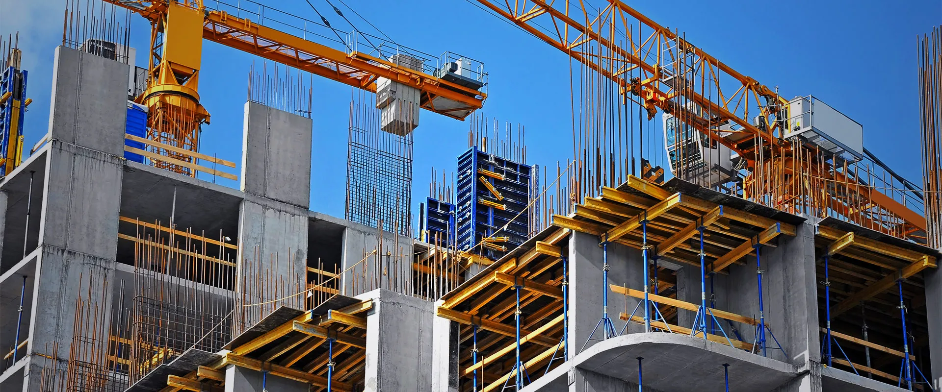 Concrete building under construction with exposed steel reinforcement bars, scaffolding, and a large yellow crane against a clear blue sky.
