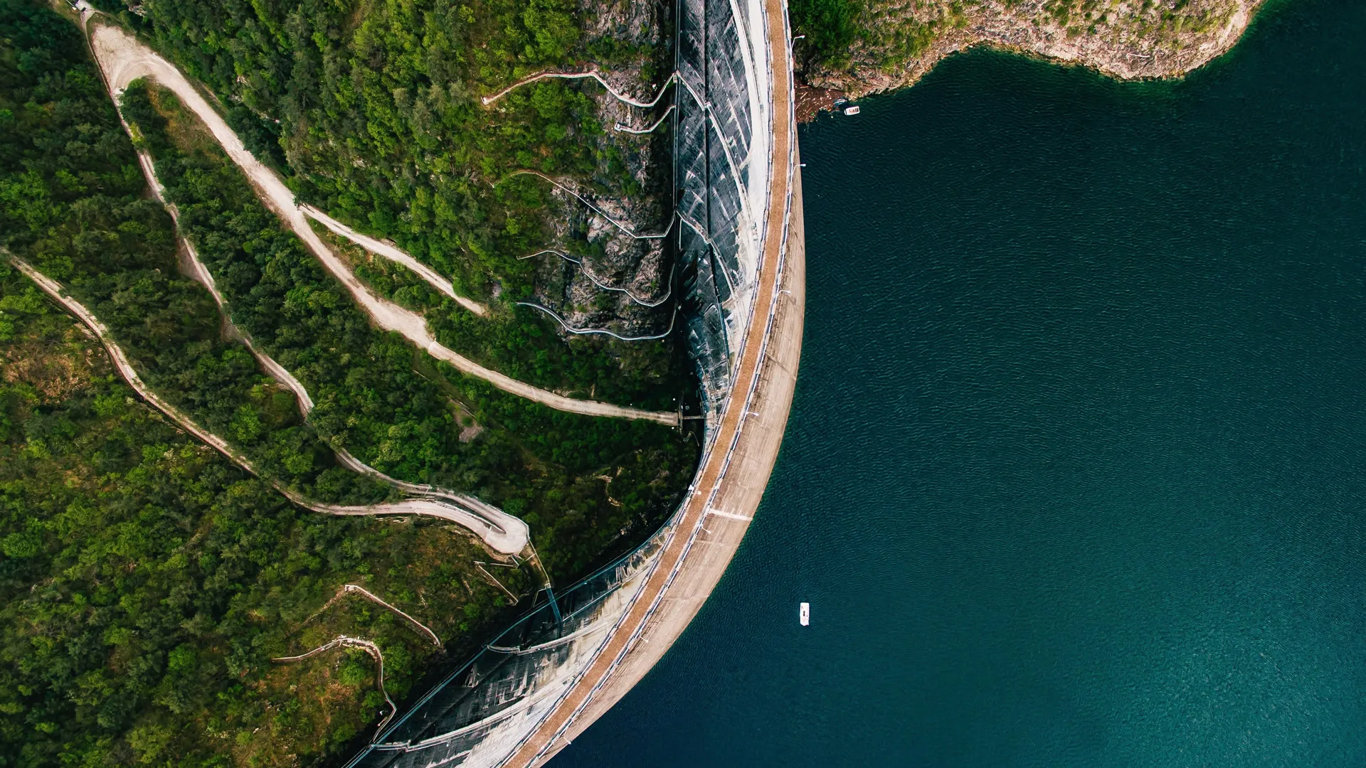 A large curved dam separates a body of blue water from a green, forested area with winding paths