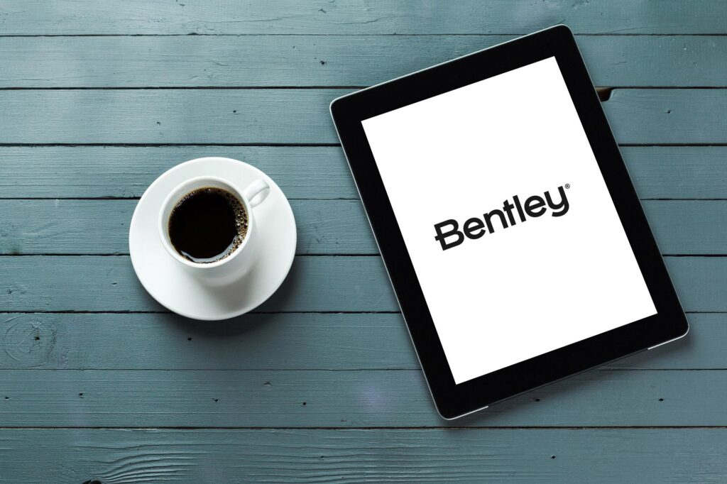 Coffee cup to the left of a tablet with the Bentley logo displaying on the tablet on blue table