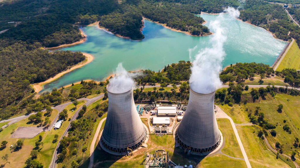 Aerial view of two large cooling towers emitting steam near a lake, surrounded by green trees and grass.