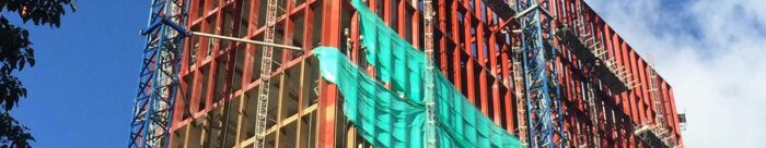 A multi-story building under construction with exposed red steel framework, blue scaffolding, and green safety netting against a partly cloudy sky.