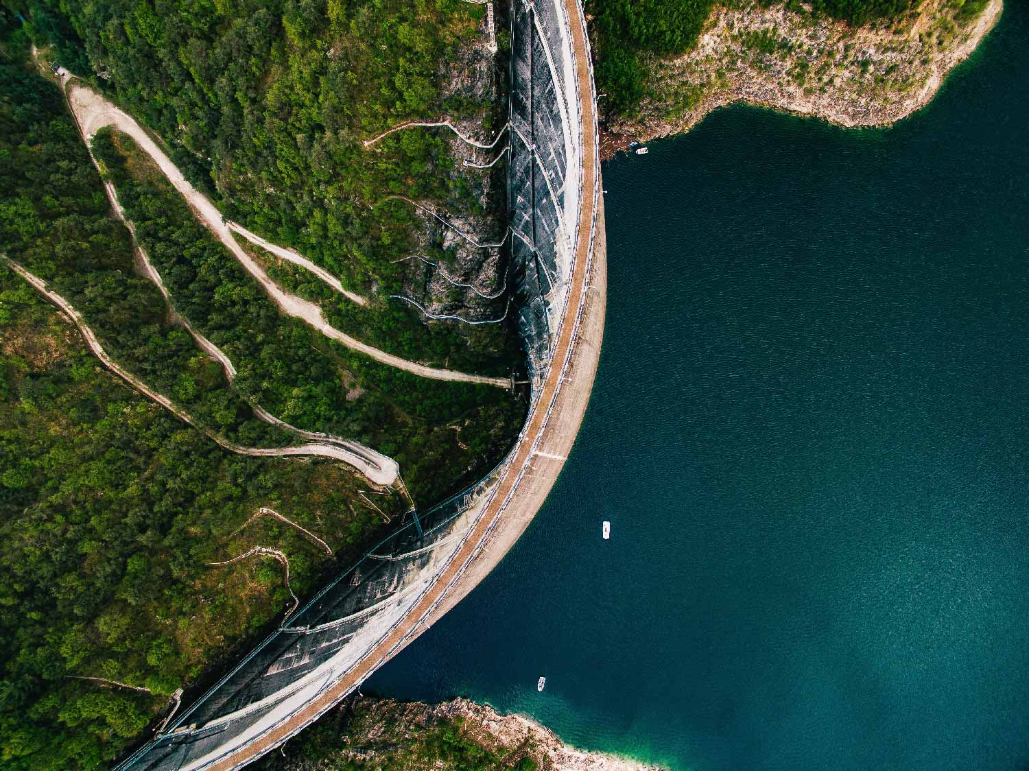 Aerial view of a large dam separating a lush green landscape from a vast body of water with small boats near the dam wall.