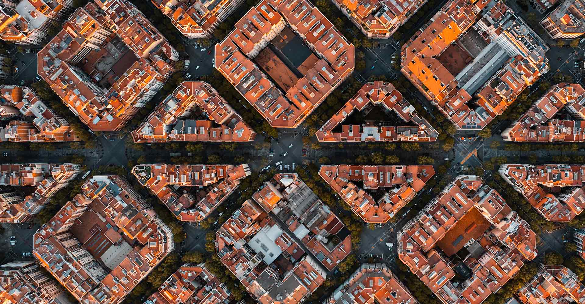 Aerial view of a cityscape with uniformly shaped, red-roofed buildings arranged in a grid pattern. Roads intersect between the blocks, and vegetation lines the streets.