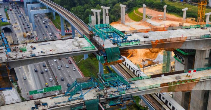 Aerial view of a highway next to a train track under construction with multiple elevated concrete structures and scaffolding. Vehicles are visible on the highway, surrounded by greenery.