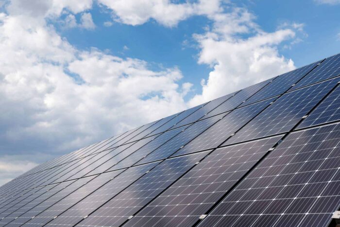 Large array of solar panels angled under a partly cloudy sky with sunlight reflecting off their surfaces.