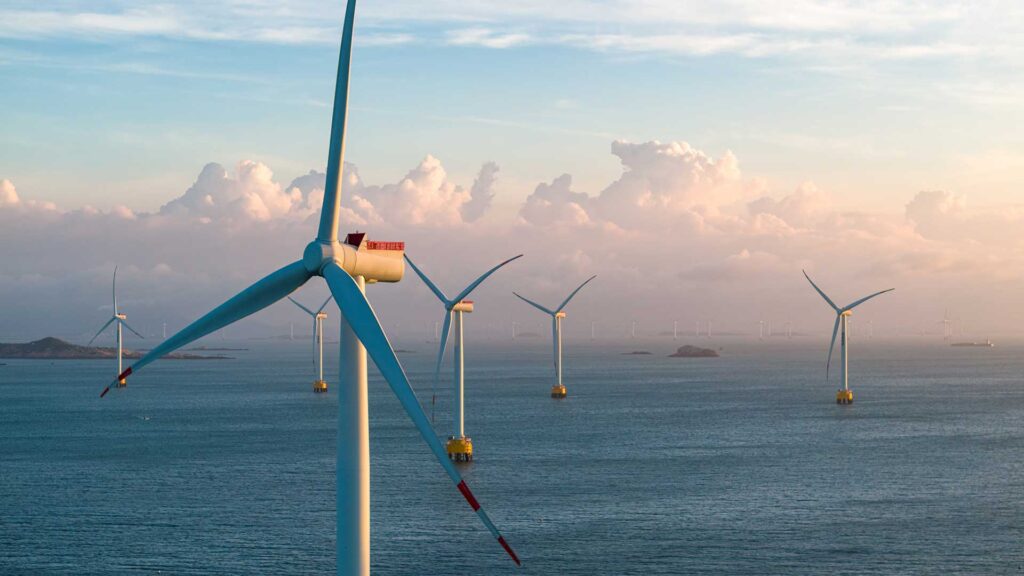 Offshore wind turbines stand in the ocean under a cloudy sky, with distant land visible on the horizon.