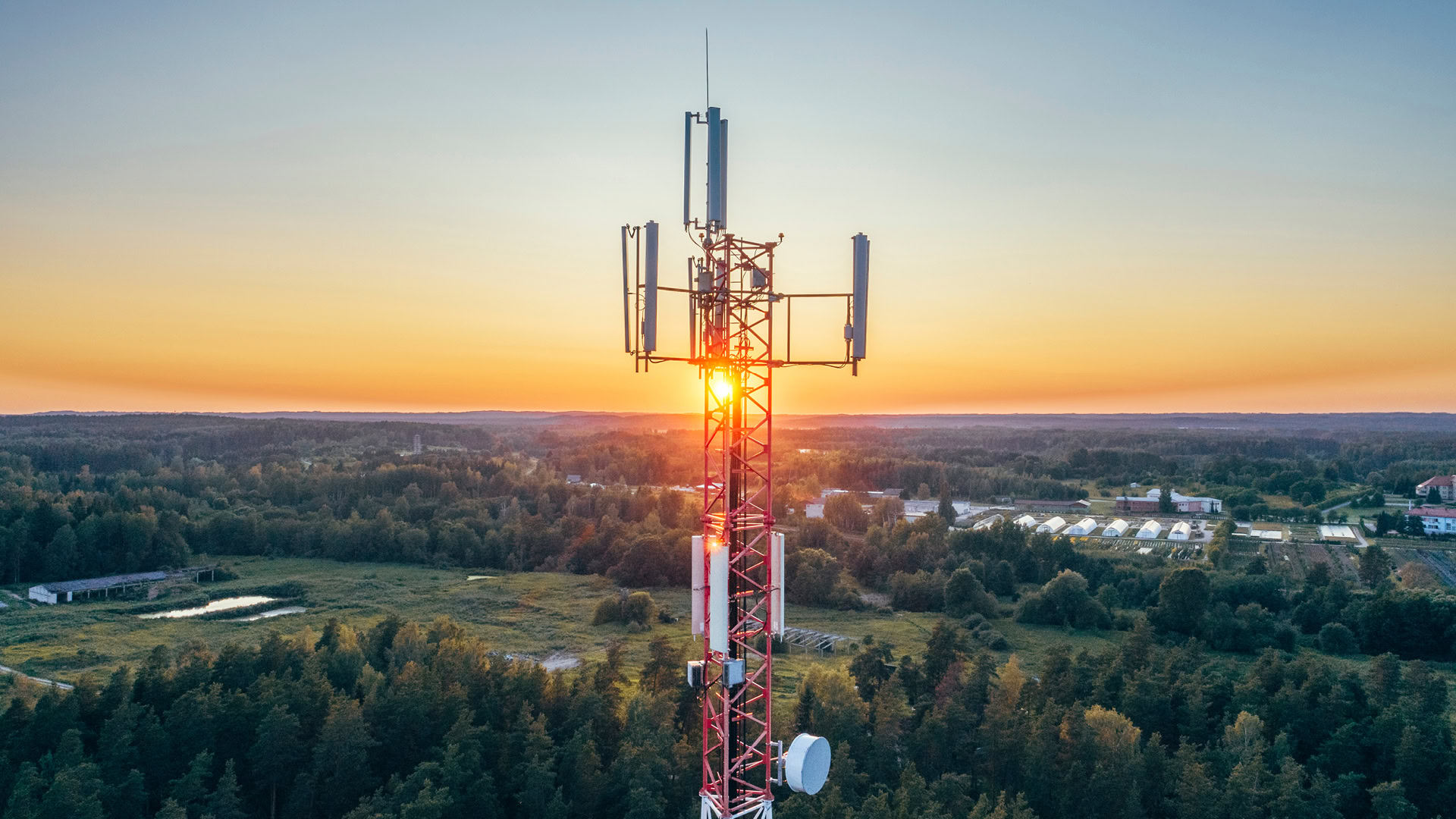 A red and white telecommunications tower stands in a forested area at sunset, with the sun visible behind the tower and a distant view of buildings and trees.