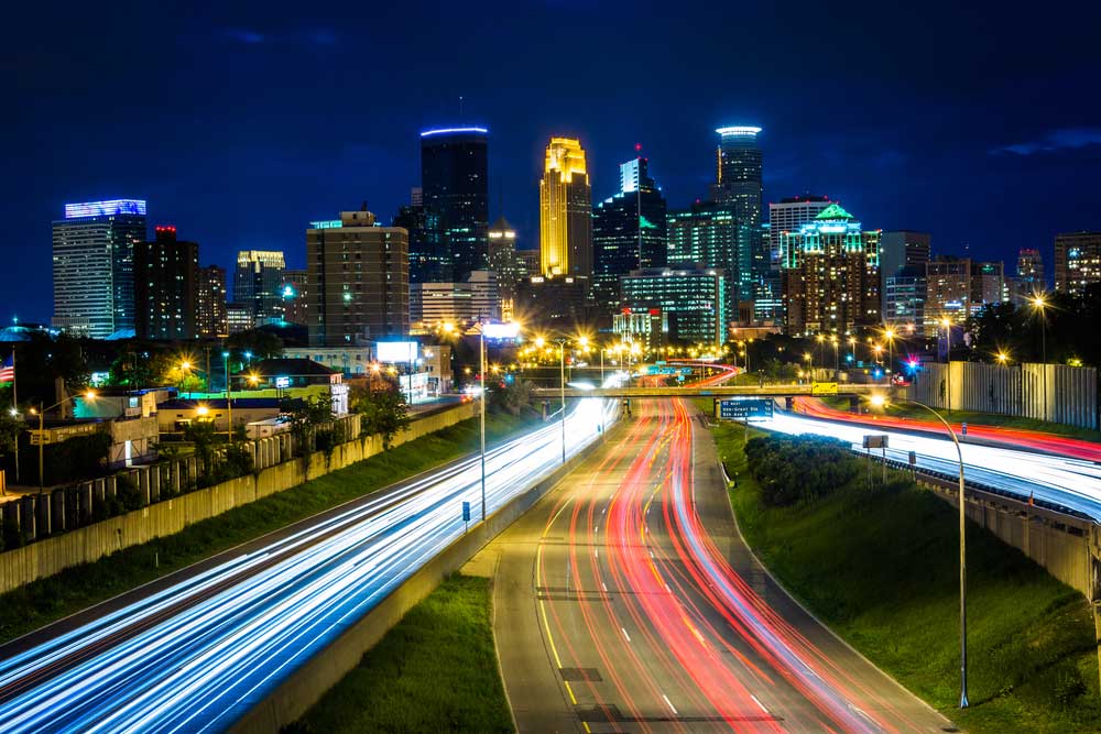 City skyline at night with illuminated skyscrapers and light trails from vehicles on a busy highway in the foreground.