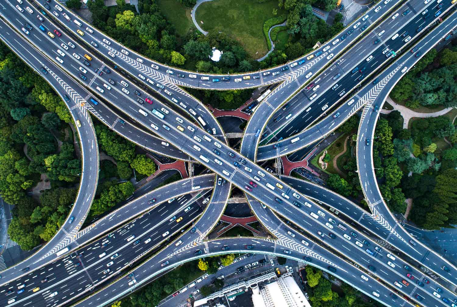 Aerial view of a complex highway interchange with multiple lanes of traffic, surrounded by green trees and some buildings.