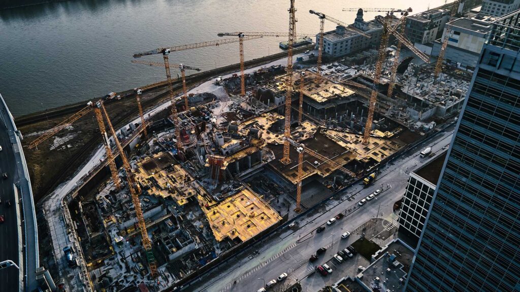 Aerial view of a large construction site with multiple cranes, buildings in various stages of development, and a river in the background.