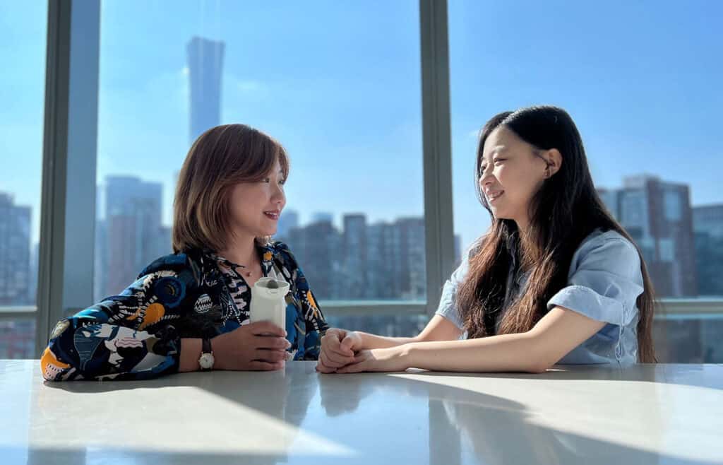 Two woman talking by a window city scape behind them