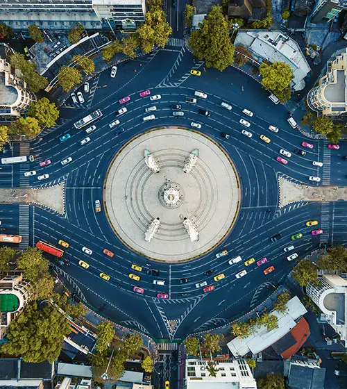 Aerial view of a large roundabout with multiple lanes of traffic, surrounding a central monument, bordered by trees and buildings.