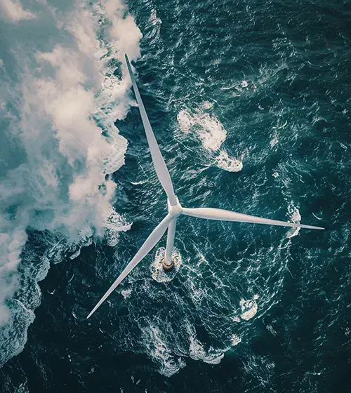 Aerial view of an offshore wind turbine in the ocean, with waves surrounding its base and water swirling nearby.