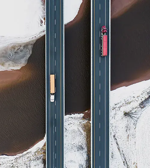 Aerial view of two parallel highways with trucks traveling in opposite directions, surrounded by snow-covered ground and brown water.