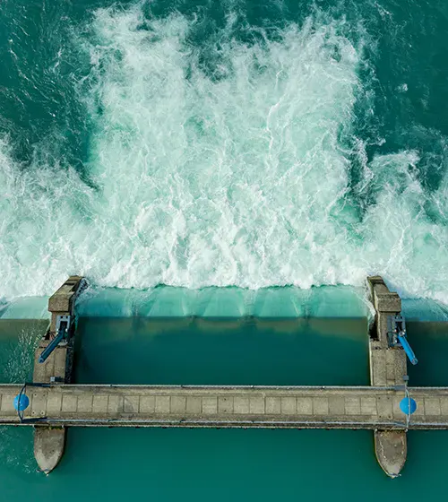Aerial view of water flowing over a dam structure, creating white turbulence as it passes through.