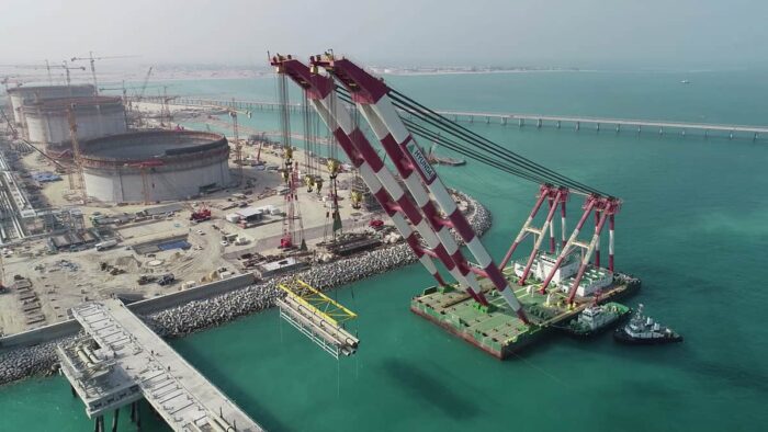 A large floating crane installs a structure in turquoise water at an industrial construction site with storage tanks, cranes, and piers in the background.