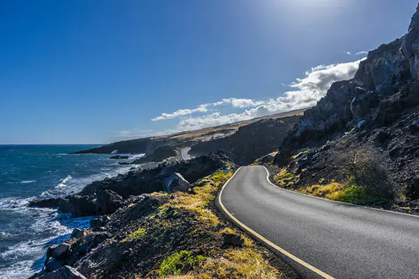 A winding asphalt road, designed with Infrastructure AI, runs along a rocky coastline with waves crashing below and cliffs rising on the right, under a clear blue sky.