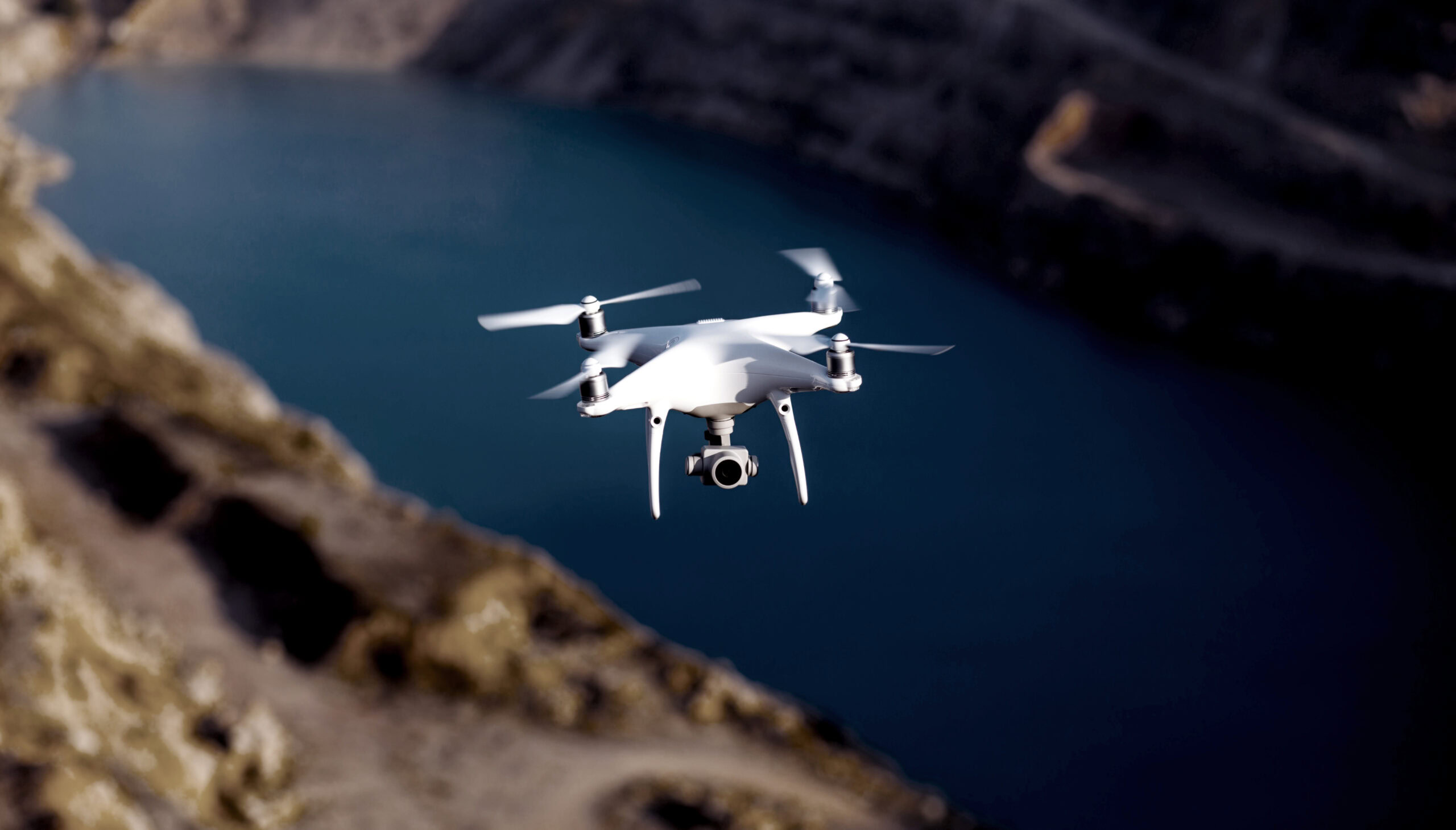 A white quadcopter drone with a camera, equipped with Infrastructure AI, hovers in the air over a blurred landscape featuring water and rocky terrain.