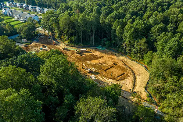 Aerial view of a construction site clearing land at the edge of a forest, with Infrastructure AI aiding the process and houses visible in the upper left background.