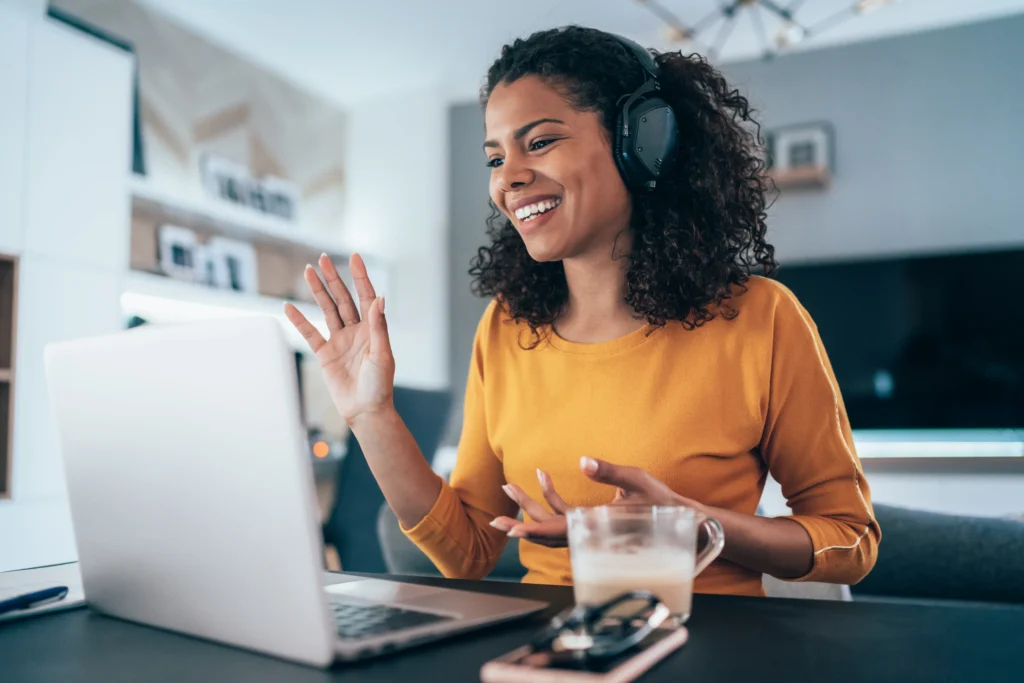 A woman wearing headphones smiles and waves at a laptop screen during a video call. She is sitting at a desk with a smartphone, a glass of beverage, and some SACS documents that hint at her offshore project in front of her.