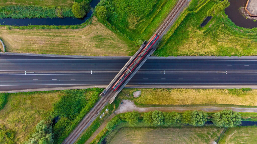 A red passenger train travels on tracks crossing over a multi-lane highway, surrounded by green fields and small waterways, viewed from above.