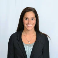 Portrait of a smiling woman with long dark hair, wearing a black blazer and a light blue top, standing against a plain white background during a webinar.