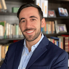 A smiling man with dark hair and a beard, dressed in a dark blazer, sitting in a room with bookshelves in the background during a webinar series.