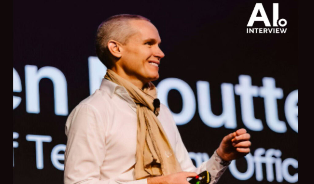 A person in a white shirt and tan scarf speaks on stage with "AI Interview" text and large projected lettering in the background, discussing infrastructure engineering and AI agents at a Bentley Systems event.
