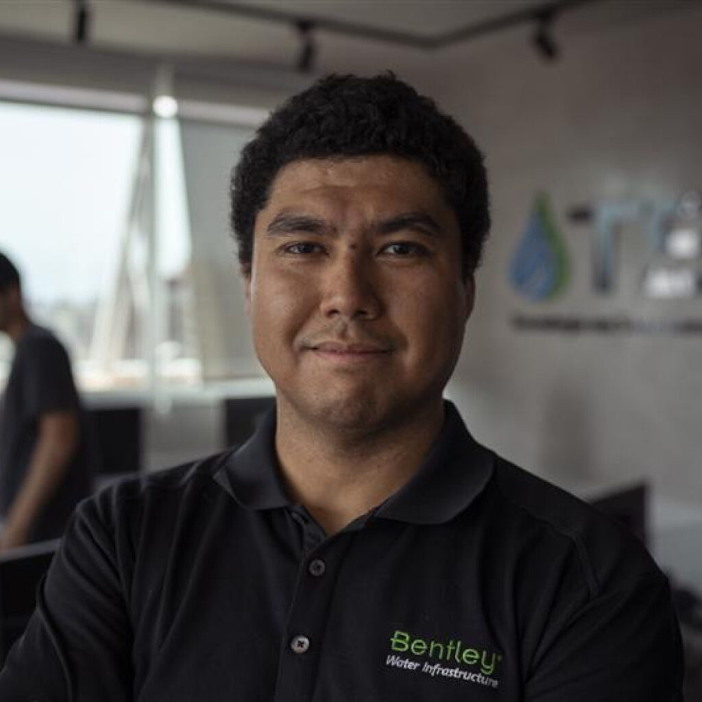 Mężczyzna ubrany w czarną koszulkę polo „Bentley Water Infrastructure” stoi w pomieszczeniu, twarzą do kamery, z rozmytym tłem biura i innym członkiem zespołu Bentley w oddali.