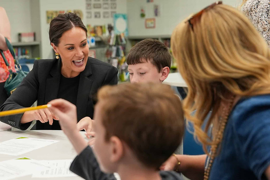 Jacqueline Coleman, in a black blazer, interacts with two children seated at a table with papers, while another adult is partially visible in the foreground—highlighting educational initiatives in Kentucky.
